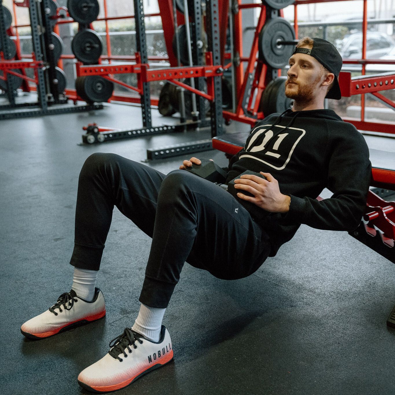 man dressed in black trousers and hoodie performing a hip thrust on a gym bench with a dumbbell on his hips, sideways to the camera