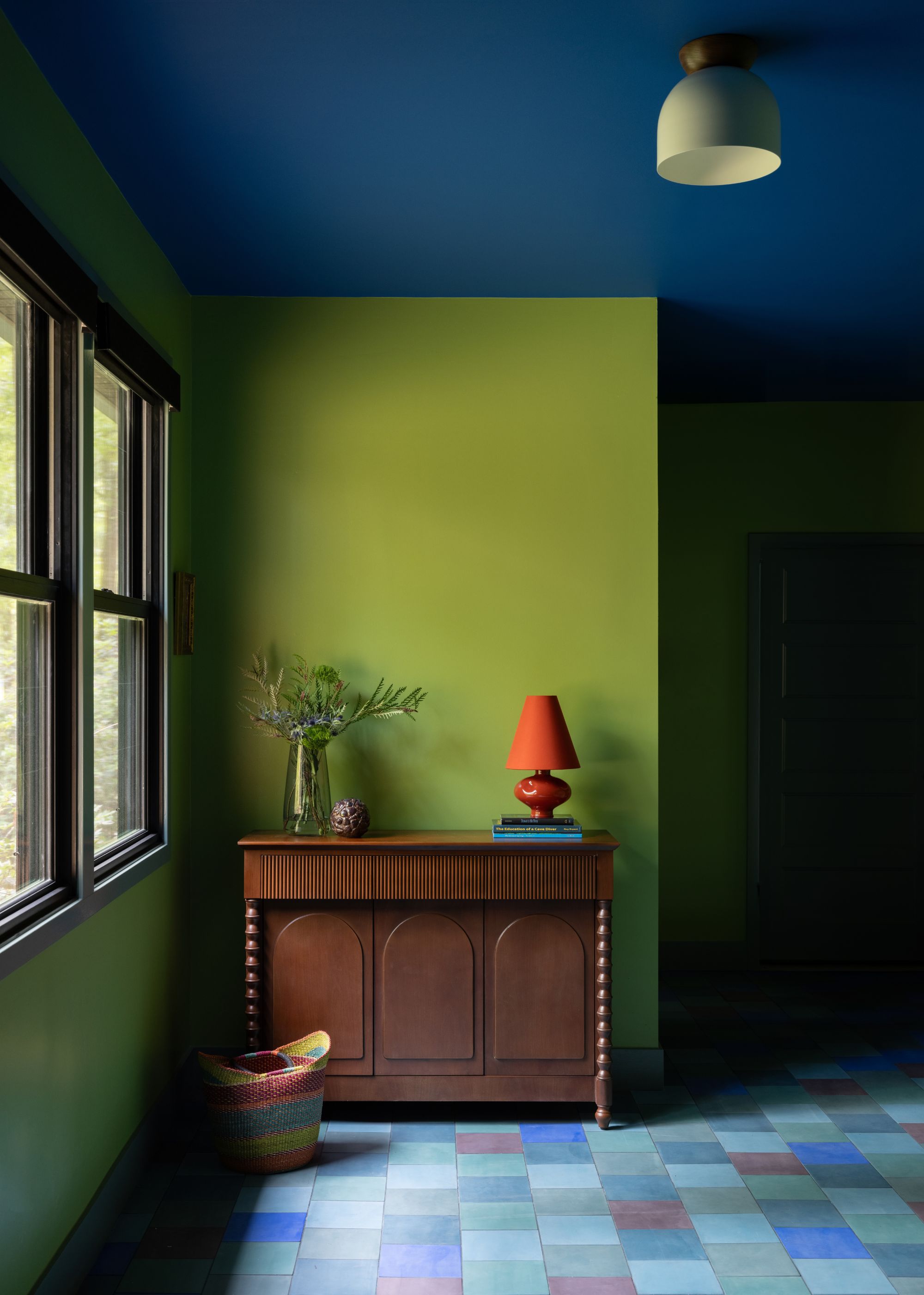 An entrance hall with lime green walls, a dark blue ceiling, square tiled flooring, and a wooden console table with a red lamp.