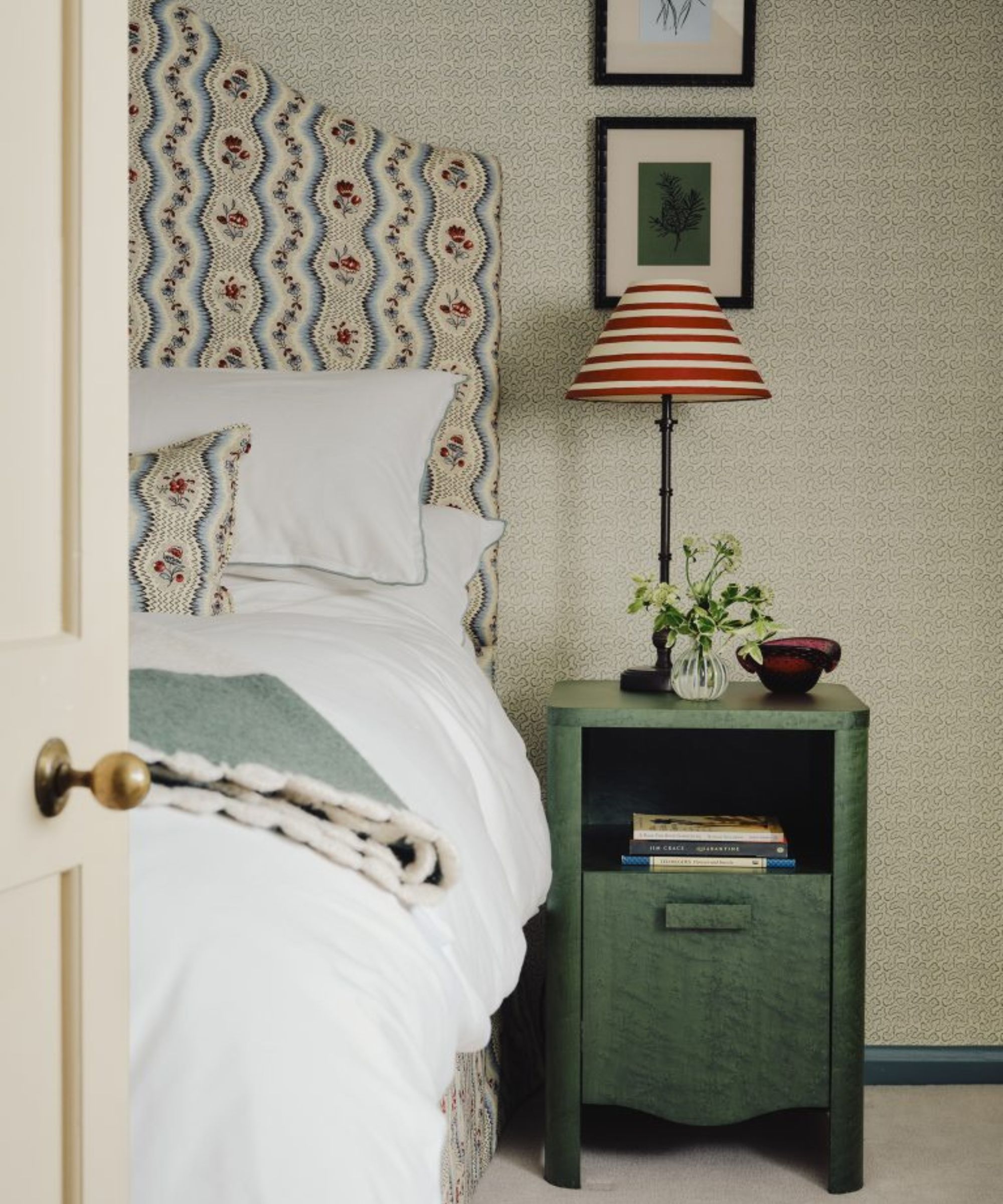 neutral small print wallpaper in a bedroom with a floral headboard and a green nightstand with a vintage lamp with red and white striped shade