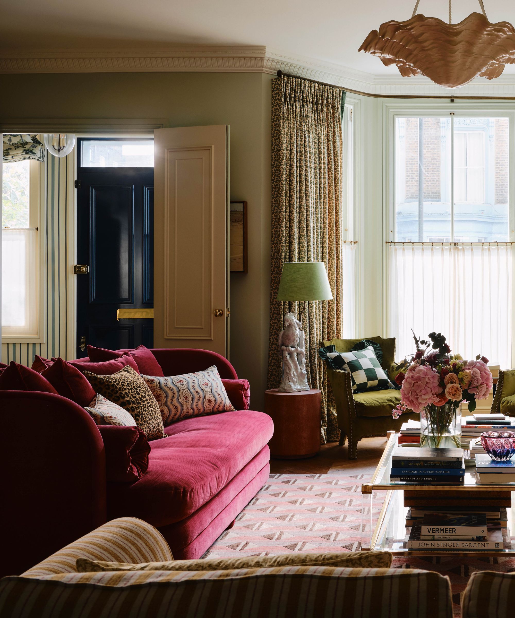 eclectic living room with a dark raspberry red velvet couch with a floral and leopard print pillows, a large bay window with cafe curtain, small green armchairs, vintage glass coffee table and a printed rug