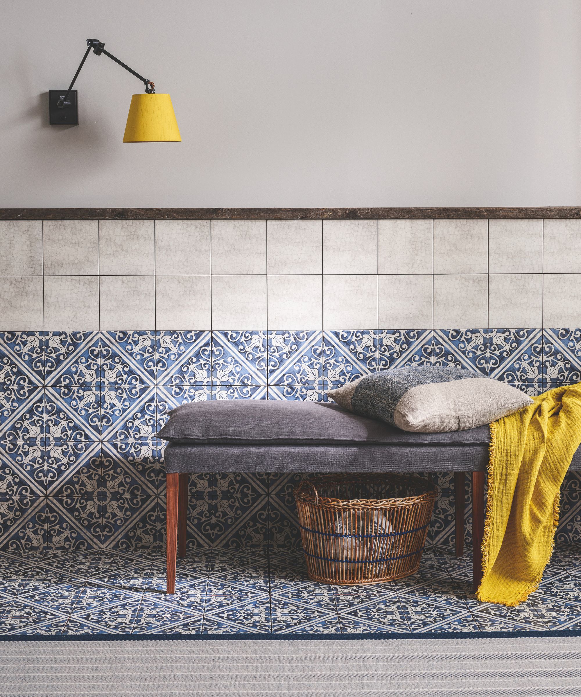 A grey upholstered bench with wooden legs against a blue and white tiled floor and decorative, patterned wall tiles. There's a yellow throw, cream and blue cushion on the bench, a wicker storage basket beneath, and a wall-mounted yellow lampshade above.
