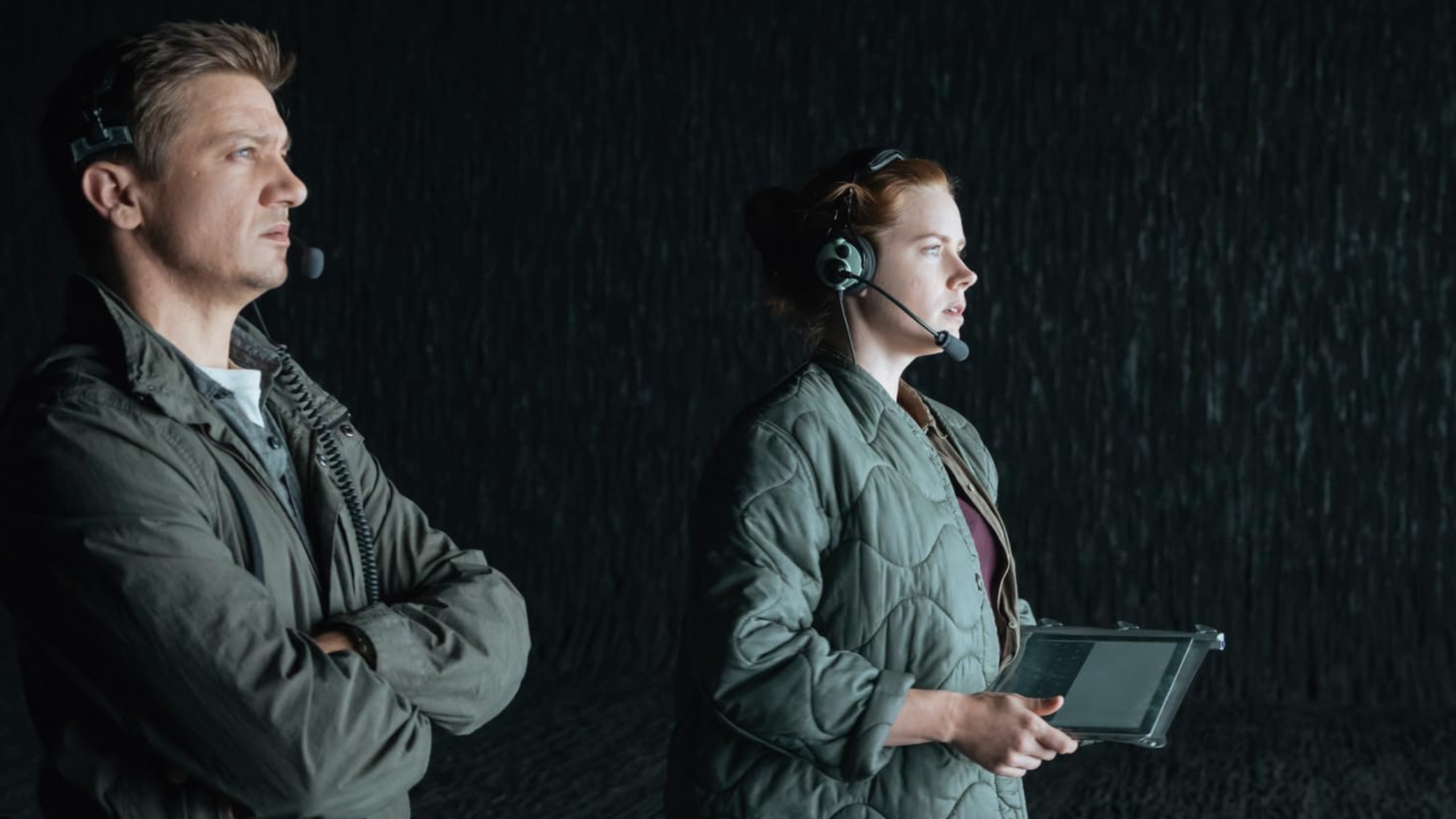 A man and a woman in outdoor clothing, staring at something off to the right of the screen. The woman is holding a tablet, and both are wearing headsets.
