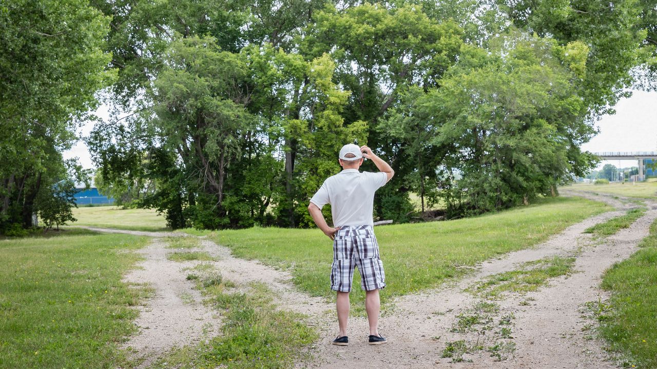 An older man stands at a fork in a road and scratches his head.