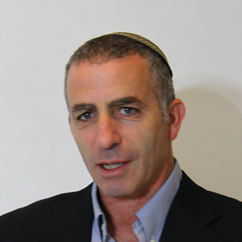 A man wearing a black blazer and a blue button up shirt with gray hair looks at the camera