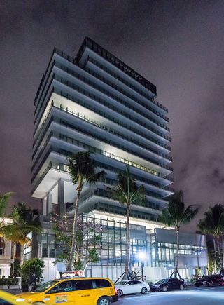 Night view of Rene Gonzalez’s crystalline tower. High-rise building with glass banisters and palm trees in front of it.