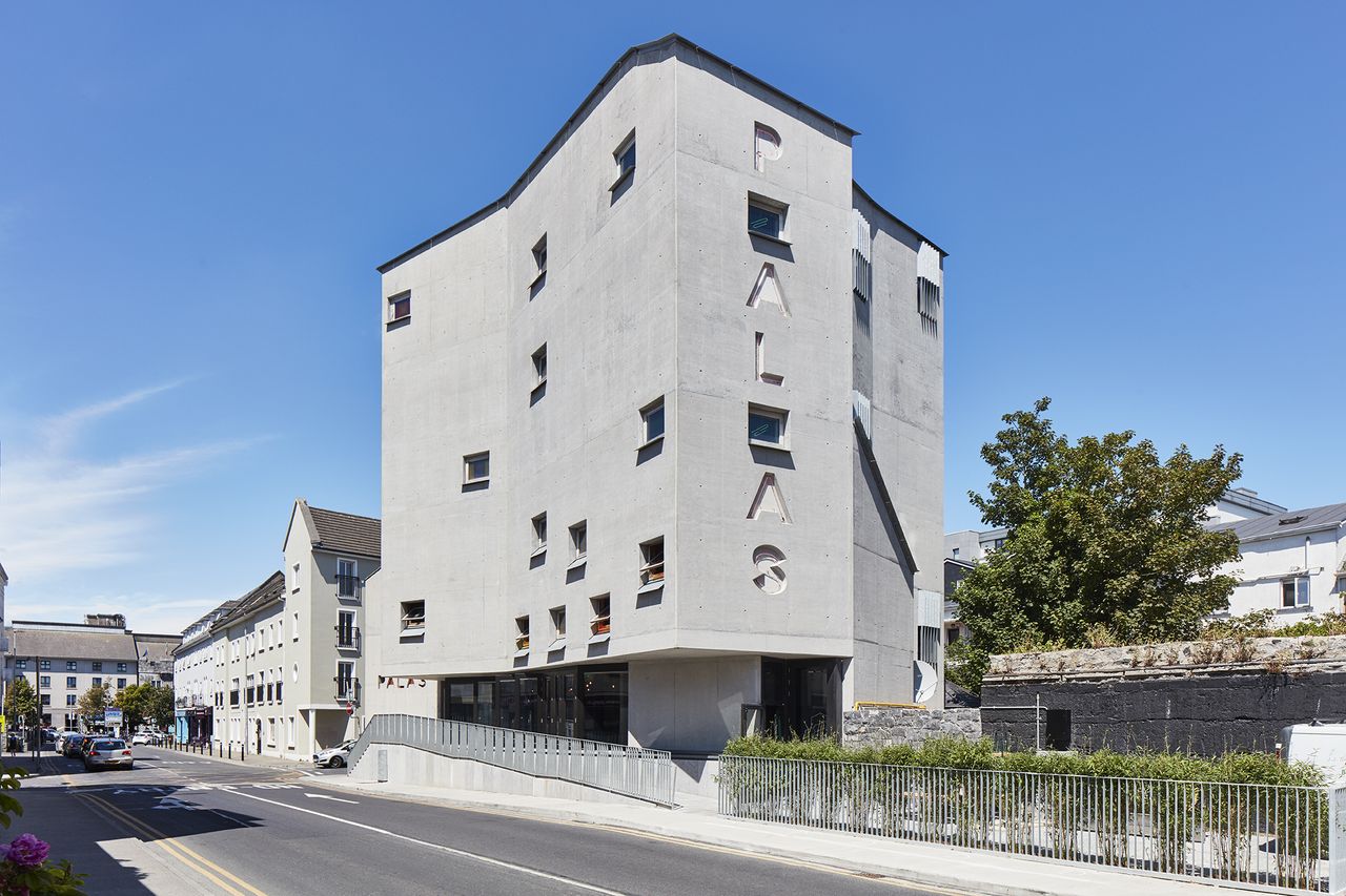 Exterior view of the grey concrete Pálás Cinema building under a clear blue sky. The building features multiple windows and the wording &#039;PÁLÁS&#039; on the side. There are residential buildings, greenery and a road nearby