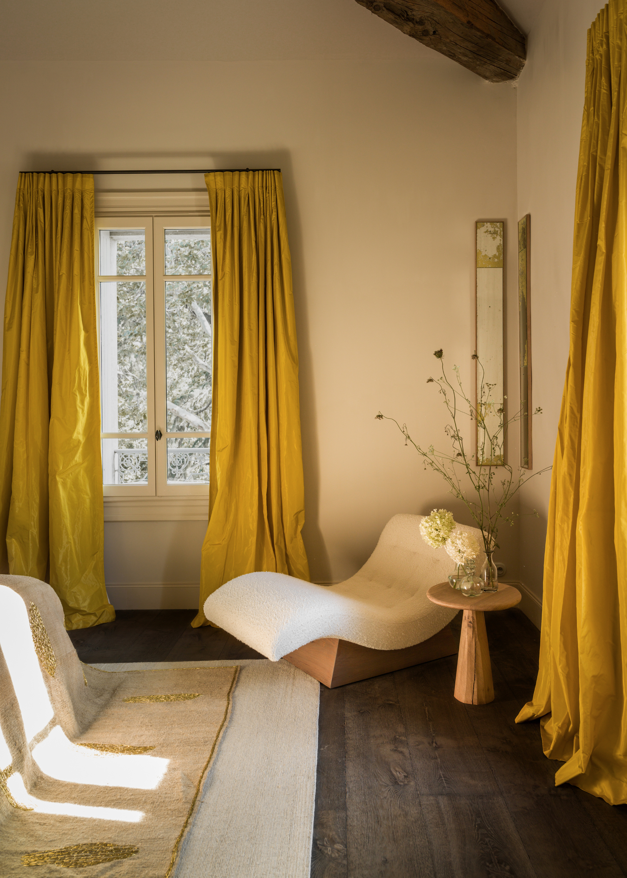 A bright bedroom with yellow silky window treatments, a white chaise lounger, a side table with vases of blooms