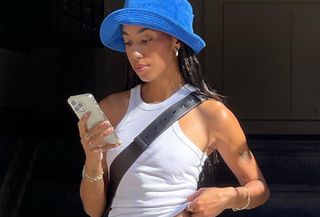 Woman wearing white tank top, blue bucket hat, and wide leg jeans shopping on phone.