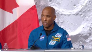 A bald man wearing a blue flight suit with the NASA meatball logo and American flag on it speaks into a microphone sitting behind a large desk with a gray background behind him.