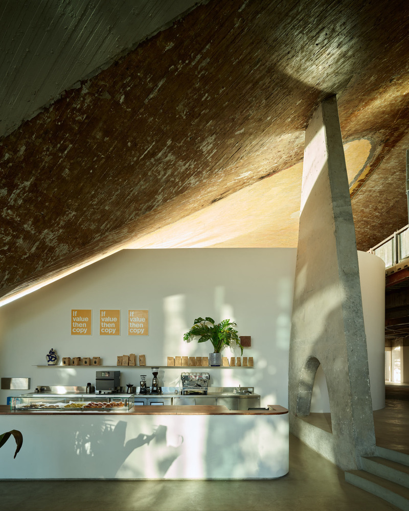 A restaurant housed inside a modernist building with exposed rock surfaces and contemporary wooden design boasts a wooden cafe bar counter decorated with chrome surfaces, stacked paper bags, plants, and more, as natural light hits its interior.