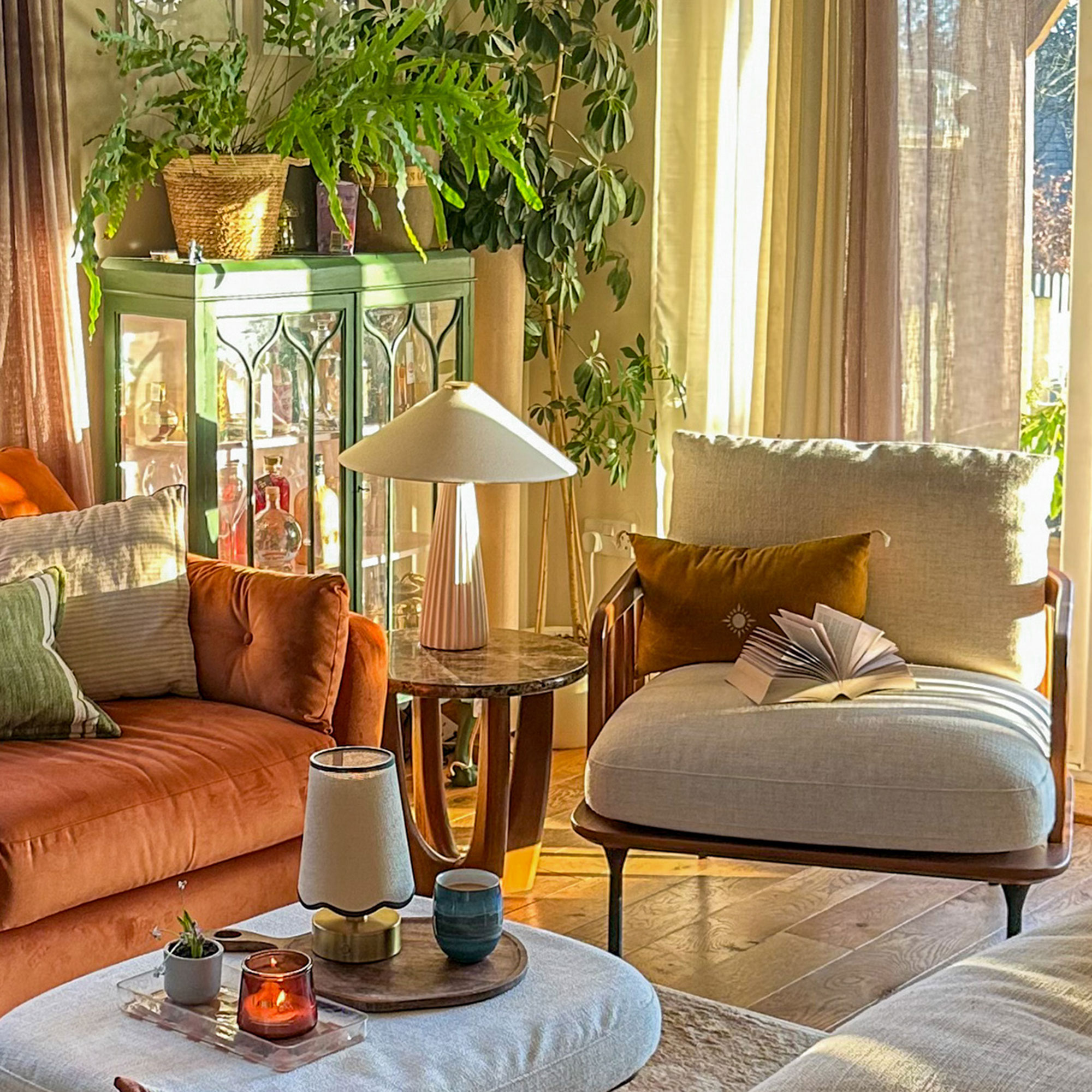 Living room with armchair, marble side table with lamp and burnt orange sofa in front of green-framed drinks cabinet topped with houseplants