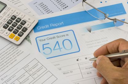 Credit report form on a desk with other paperwork. There are also a pen, glasses and a calculator on the desk. Hand is holding pen