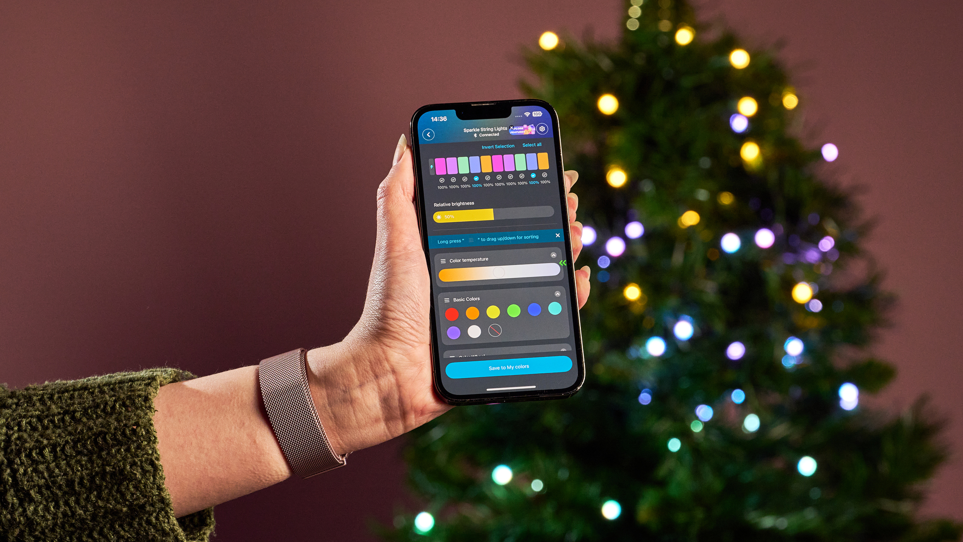 A woman holds a smartphone showing the Govee app. In the background, the Govee Sparkle String Lights are glowing multiple colors on a Christmas tree.