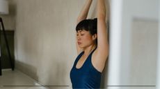 Woman practising wall Pilates at home with arms stretched out above her head and eyes closed