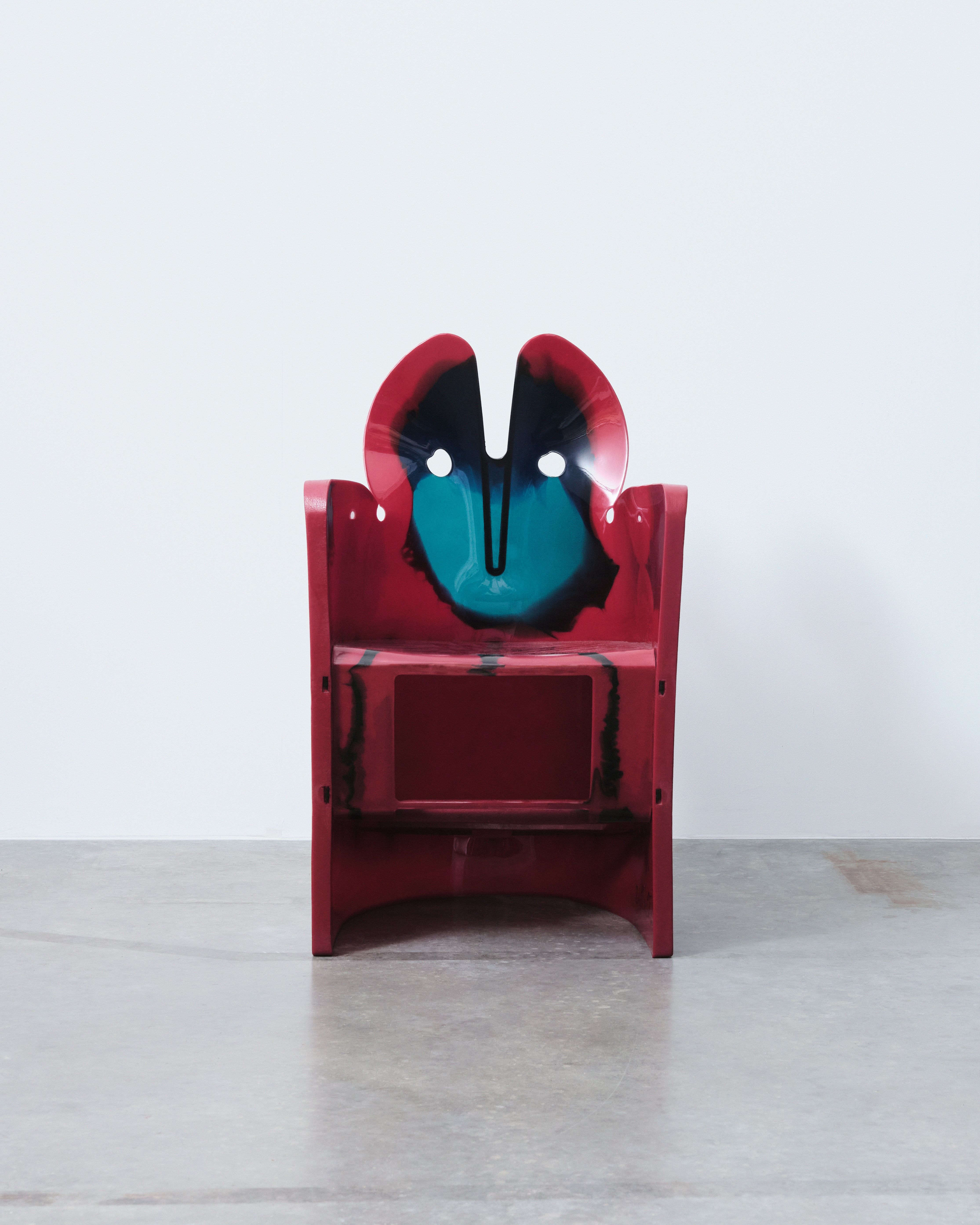A resin, red, blue, and black-painted sculptural chair with two holes looking like animal eyes and ears like those of a bunny sits in an industrial site.