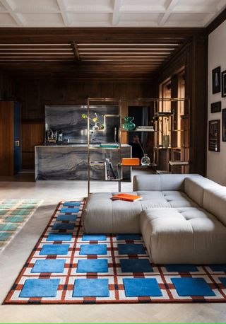 Red and blue rug in a mid-century modern living room. There are white floors, wooden walls, and a large, off-white sectional couch. A marble bar is in the background.