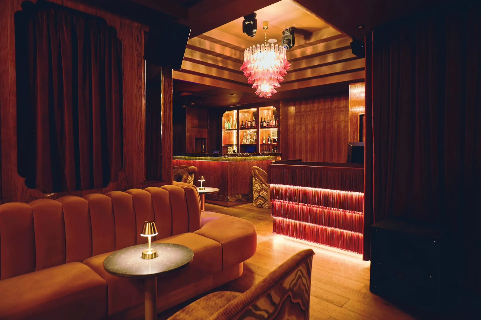A 1970s-style, lavish salon with velvety, ochre banquettes, murano glass chandeliers, reflective surfaces, red curtains, and brass table lamps, shown alongisde a back-lit, glamorous, sleek bar.