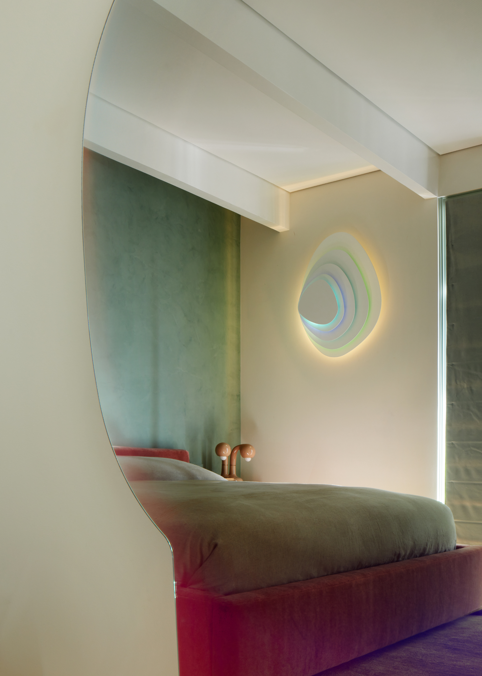 A mirror with a reflection of a magenta bed frame, a green bedscape, a wood lamp, and a stacked lighting feature