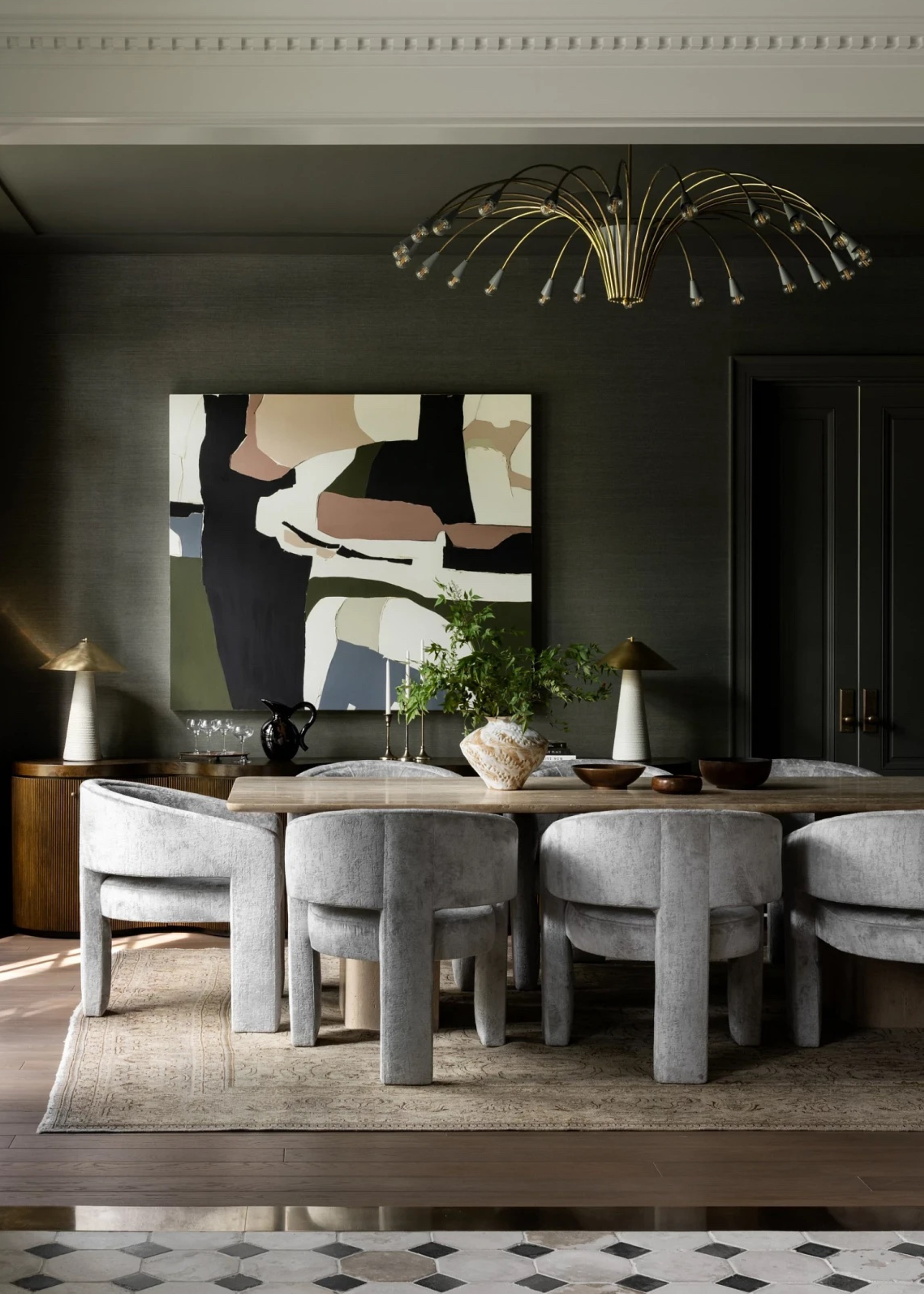 A dark green dining room with a metal chandelier, a colorful painting, a wood table with a vase and some bowls, and a set of round-backed gray chairs