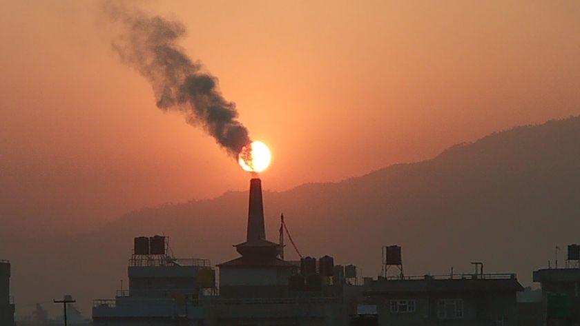 Smoke billows from a tall tower silhouetted by a rising orange/yellow sun behind the city