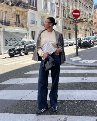 French fashion influencer Lena Farl poses on a Paris crosswalk wearing eyeglasses, a low bun, teardrop earrings, a houndstooth print blazer, cream sweatshirt, mini black bag, dark-wash jeans, and Samba sneakers.