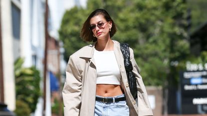 Hailey Bieber walking in the LA sunshine wearing a trench coat over a white crop top and denim mini skirt