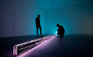 A man and a woman are standing next to the neon art installation in a dark-lit room. The installation is in a shape of a long beam with electronic neon writing on it.