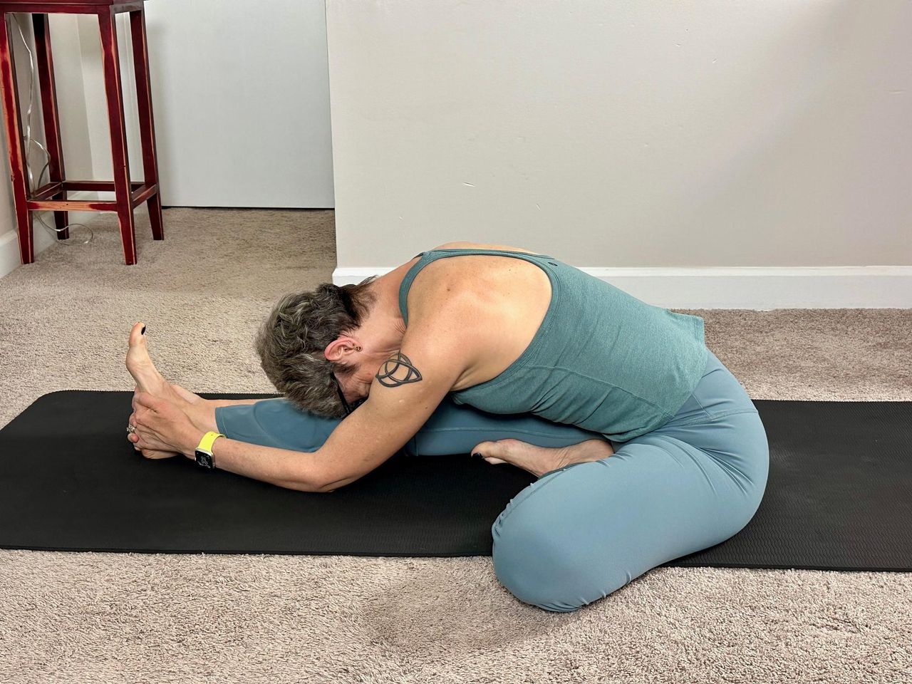 woman performing a yoga stretch sitting on the floor with one leg extended and the other bent in a living room on a black yoga mat