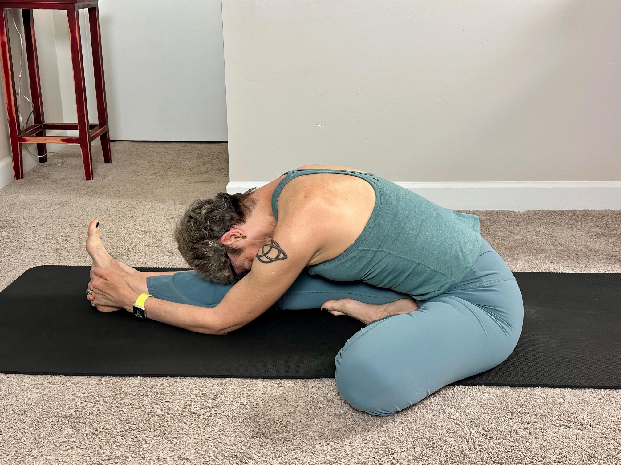 woman performing a yoga stretch sitting on the floor with one leg extended and the other bent in a living room on a black yoga mat