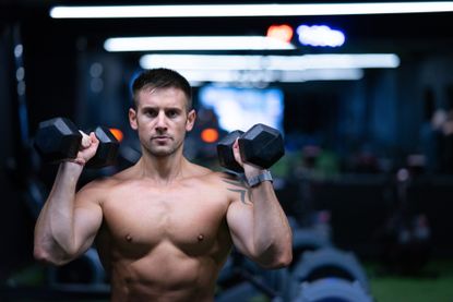 How to master shoulder press: Fitness personality Alex Crockford holding two dumbbells at shoulder height in the gym