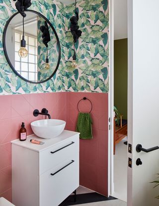 Pink wall tiles meet botanical wallpaper for a playful look in this modern bathroom, complete with round wall mirror, and white vanity unit.