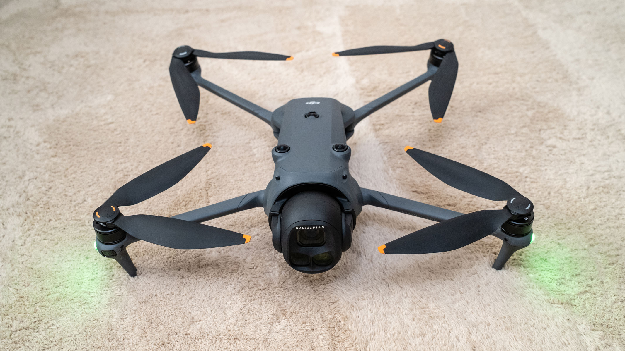 A grey drone has four propellers extending out of each corner of the central body with green lights shining from the front two propellers on a beige carpet.