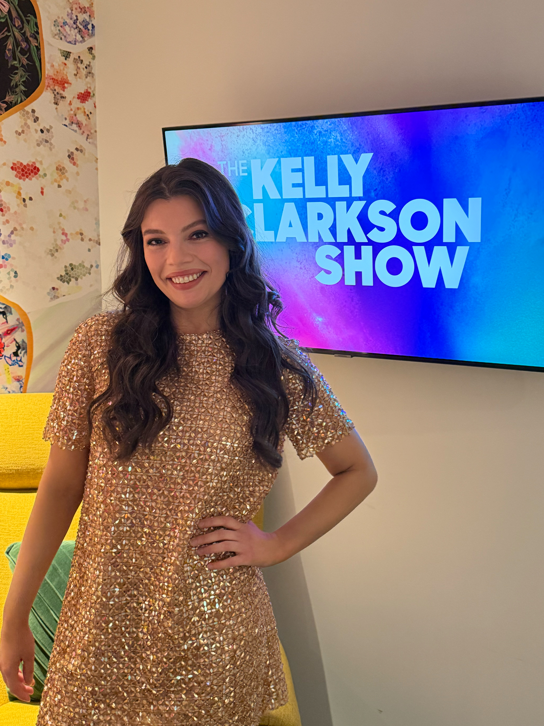 Author Alex Aster poses on the Kelly Clarkson Show wearing a gold sequin short-sleeve minidress.