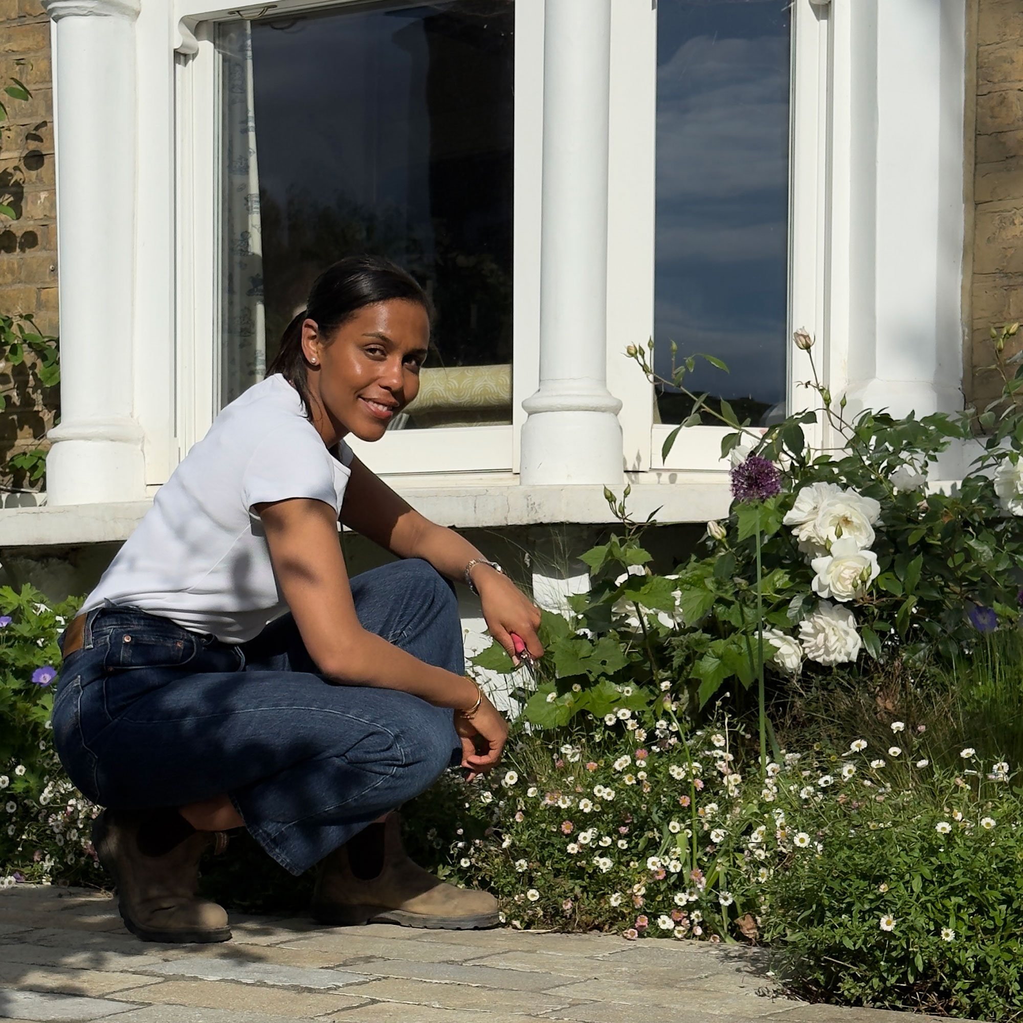 Woman kneeling outside in front of bay window looking at flowering plants