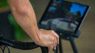 A close up of a woman's hand, which is holding the shifter of a bike while she cycles indoors, with the Rouvy app showing on a tablet in the background