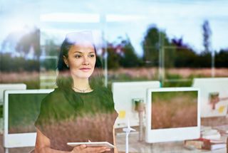 A business woman stood in a room of computers looking out of a window with a field and trees reflected on the glass