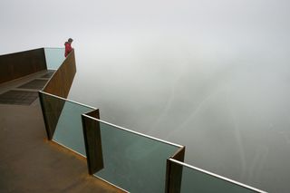 A rusted steel viewing platform with glass barriers surrounded by a white fog. There is a person in a red jacket standing on the platform looking out