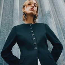 Young blonde woman wearing tailored button-up blazer, stood by steel wall.
