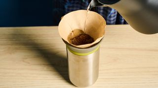 Wacaco Cuppamoka, a stainless steel coffee pour-over brewer, photographed against a blue background, with brown filters and a sippy lid