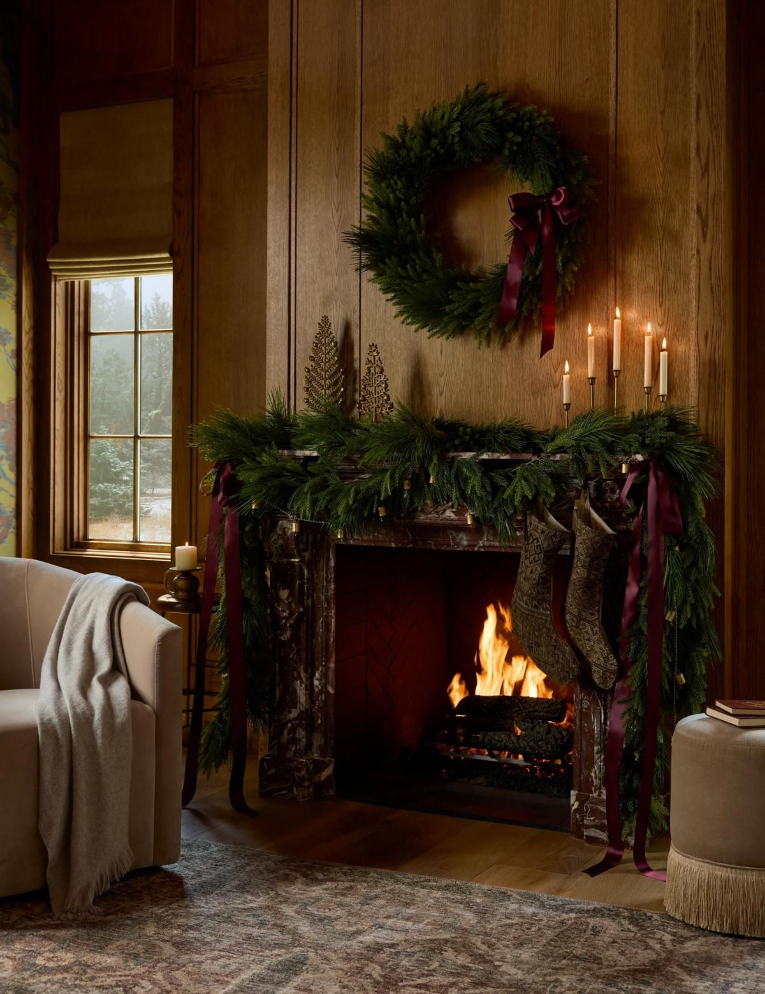 Cozy, festive living room featuring a roaring fireplace styled with garland and a Christmas wreath overhead. A sitting chair and fringed ottoman flank the wood-paneled scene.