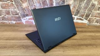 Top cover of the MSI Prestige 16 AI Evo (B2HM) business laptop on a wooden desk, in front of a brick wall.