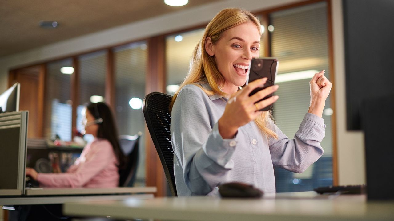 Woman sitting in an office at her desk, holding a cell phone and looking excited and happy