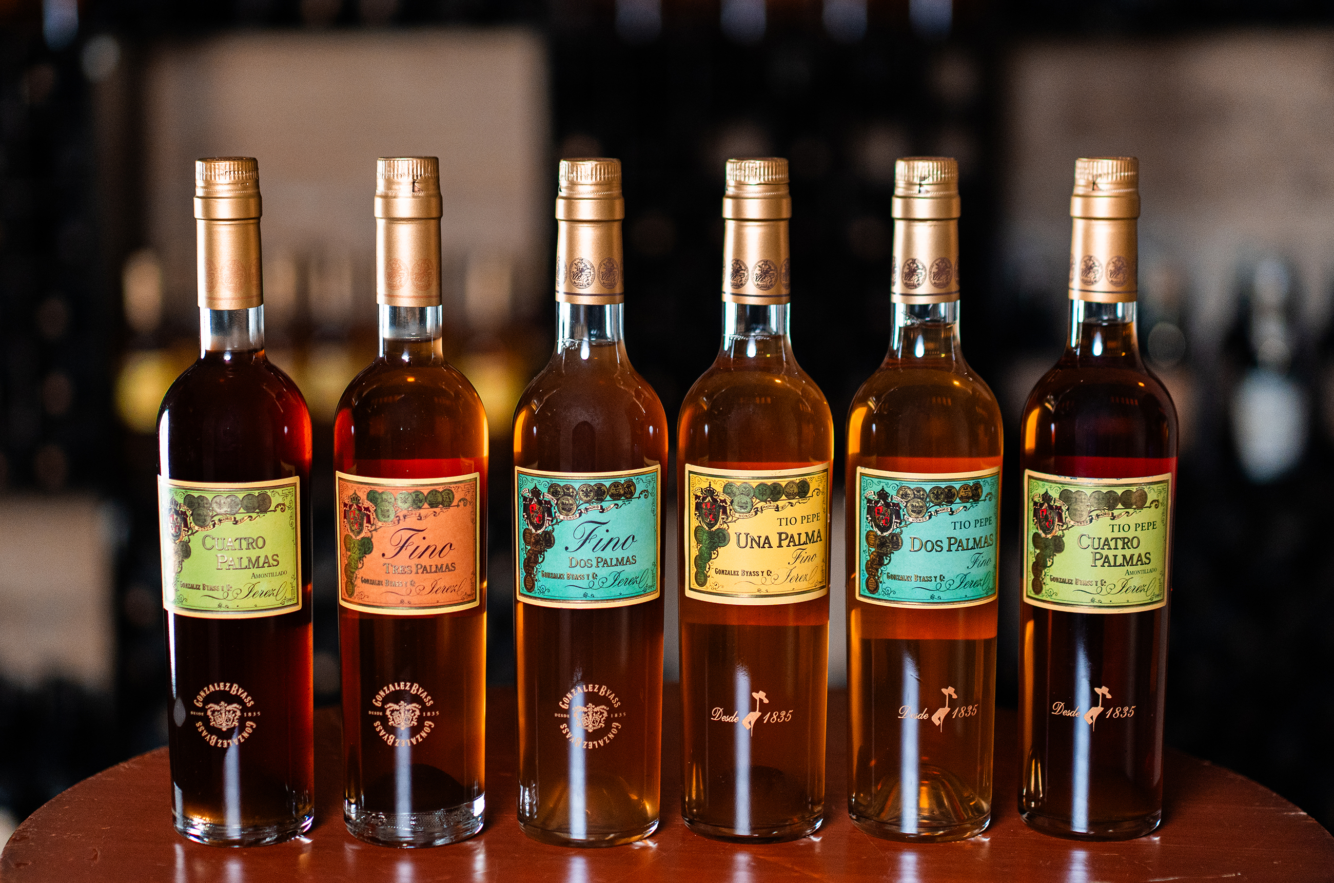 Six bottles of sherry on a table, all with different brightly coloured labels, saying 'Palmas' 