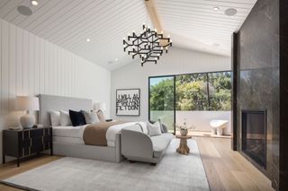 bedroom with with pale wood floor white wood cladding on walls and ceiling black marble wall grey sofa and bedhead and black chandelier and glass doors to deck