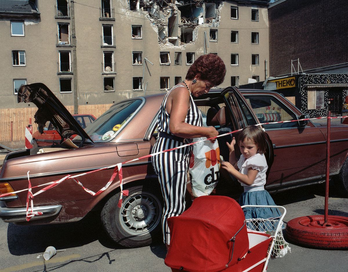 In 1988 Cologne, a woman in a black and white striped jumpsuit unpacks shopping bags beside a car and a young girl, a bomb-damaged apartment block looming in th