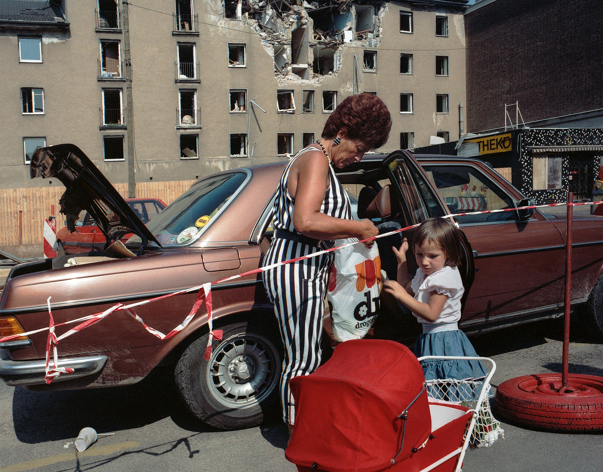 In 1988 Cologne, a woman in a black and white striped jumpsuit unpacks shopping bags beside a car and a young girl, a bomb-damaged apartment block looming in the background.