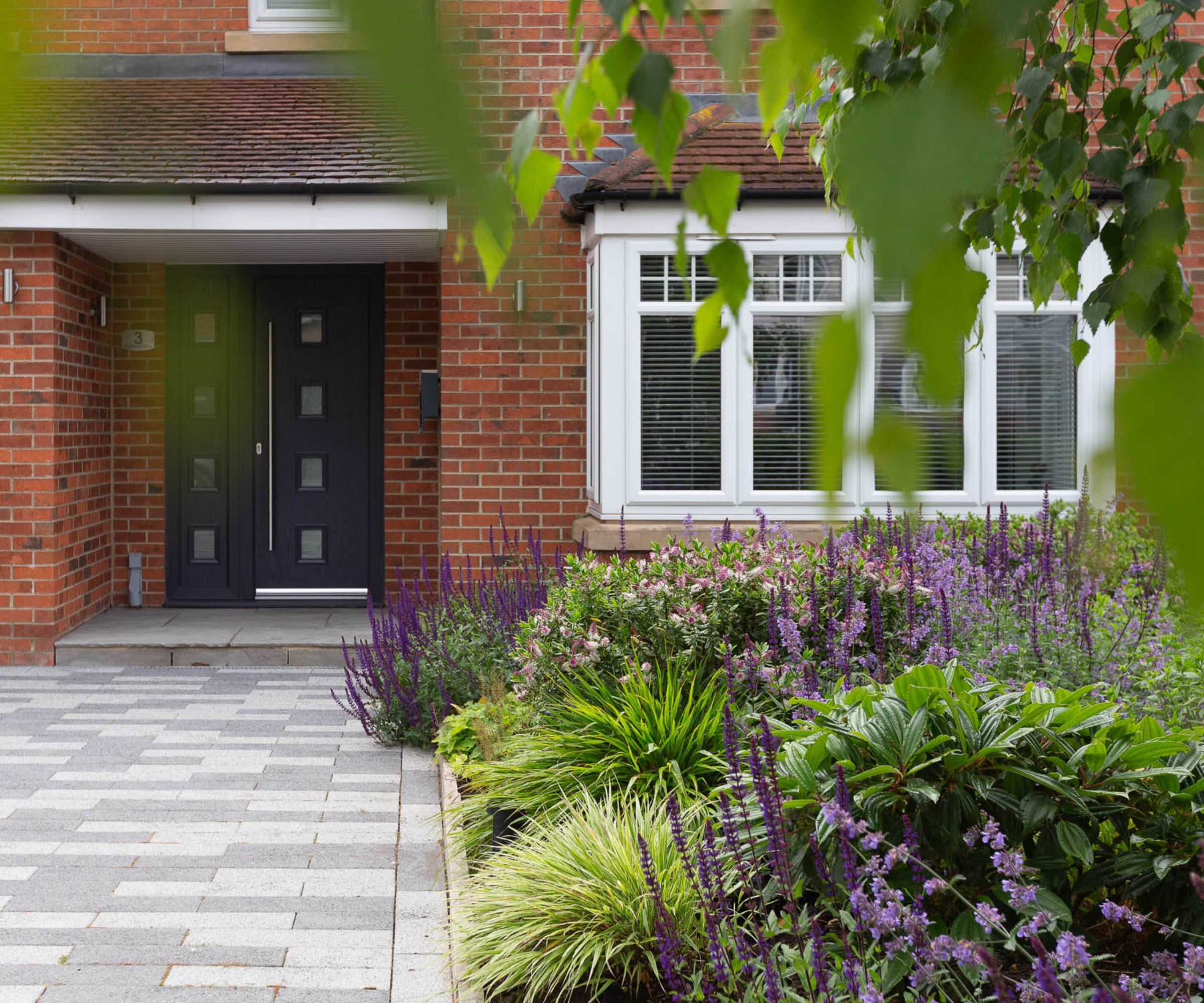 brick house with grey paved driveway and planted garden to one side with lots of green foliage, a tree and purple plants