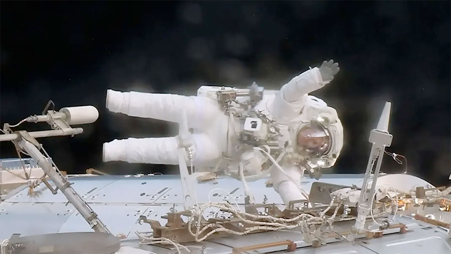 an astronaut in a white spacesuit is seen waving while positioned horizontal to the point of the view of the camera during a spacewalk outside a space station