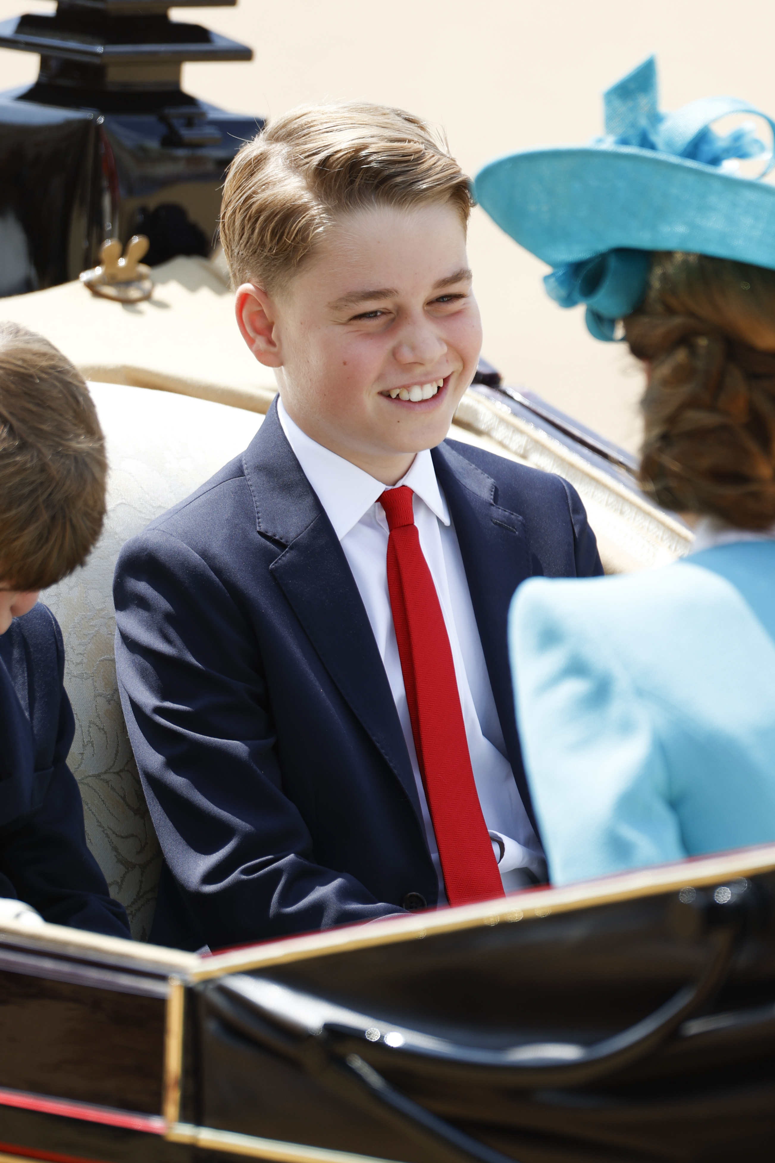 Prince George wearing a blue suit and red tie riding in a carriage