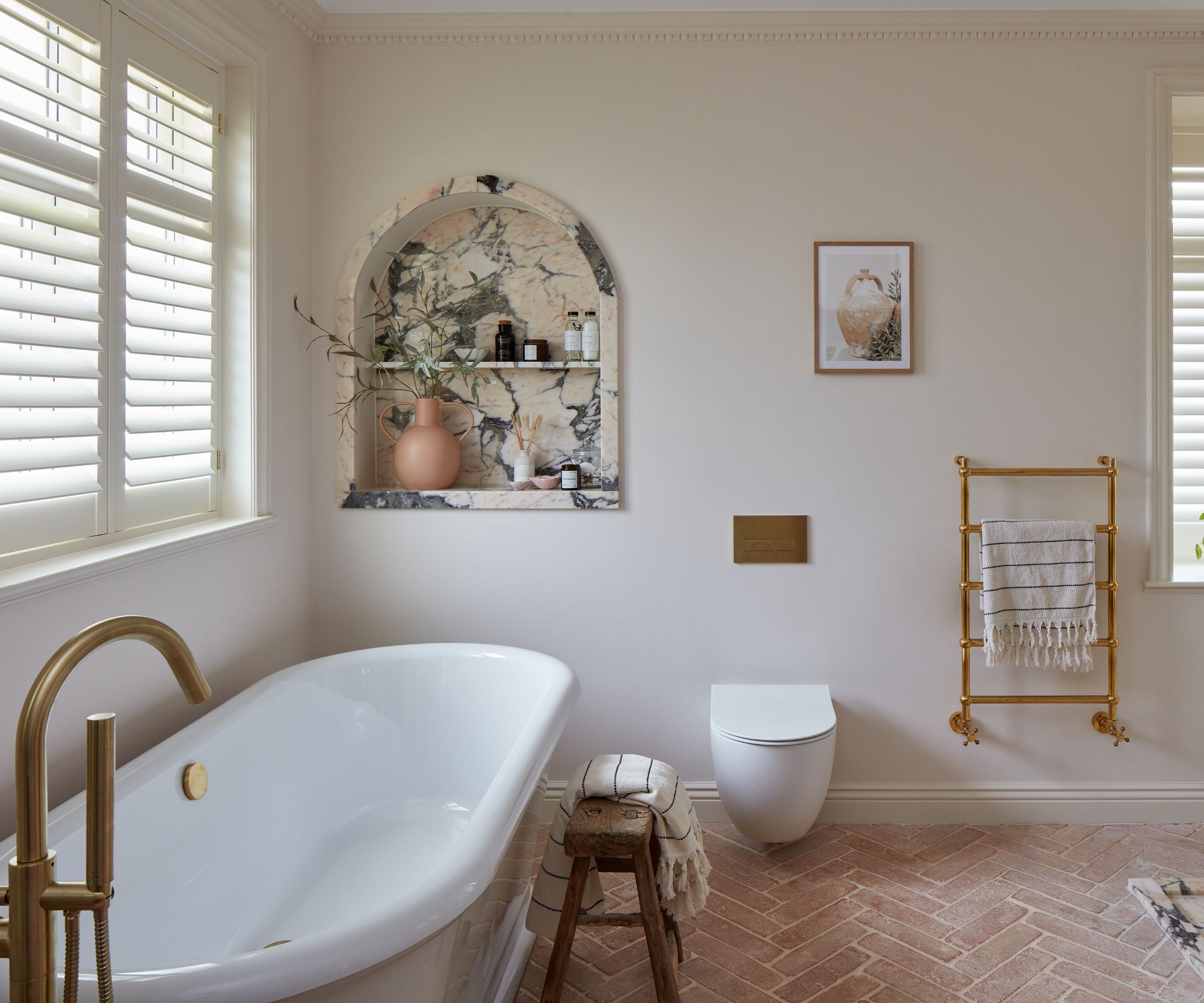 A Mediterranean bathroom with terracotta floor tiles, a freestanding bath, brass hardware, and a marble recess in the wall for decor